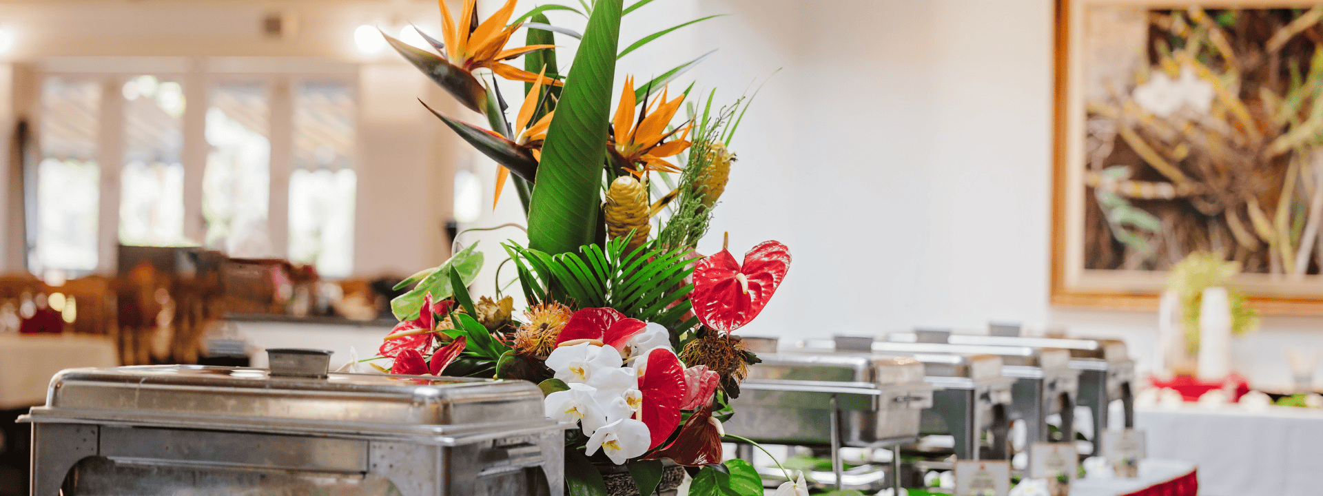 A vibrant floral arrangement featuring tropical flowers, with buffet trays in a restaurant setting.
